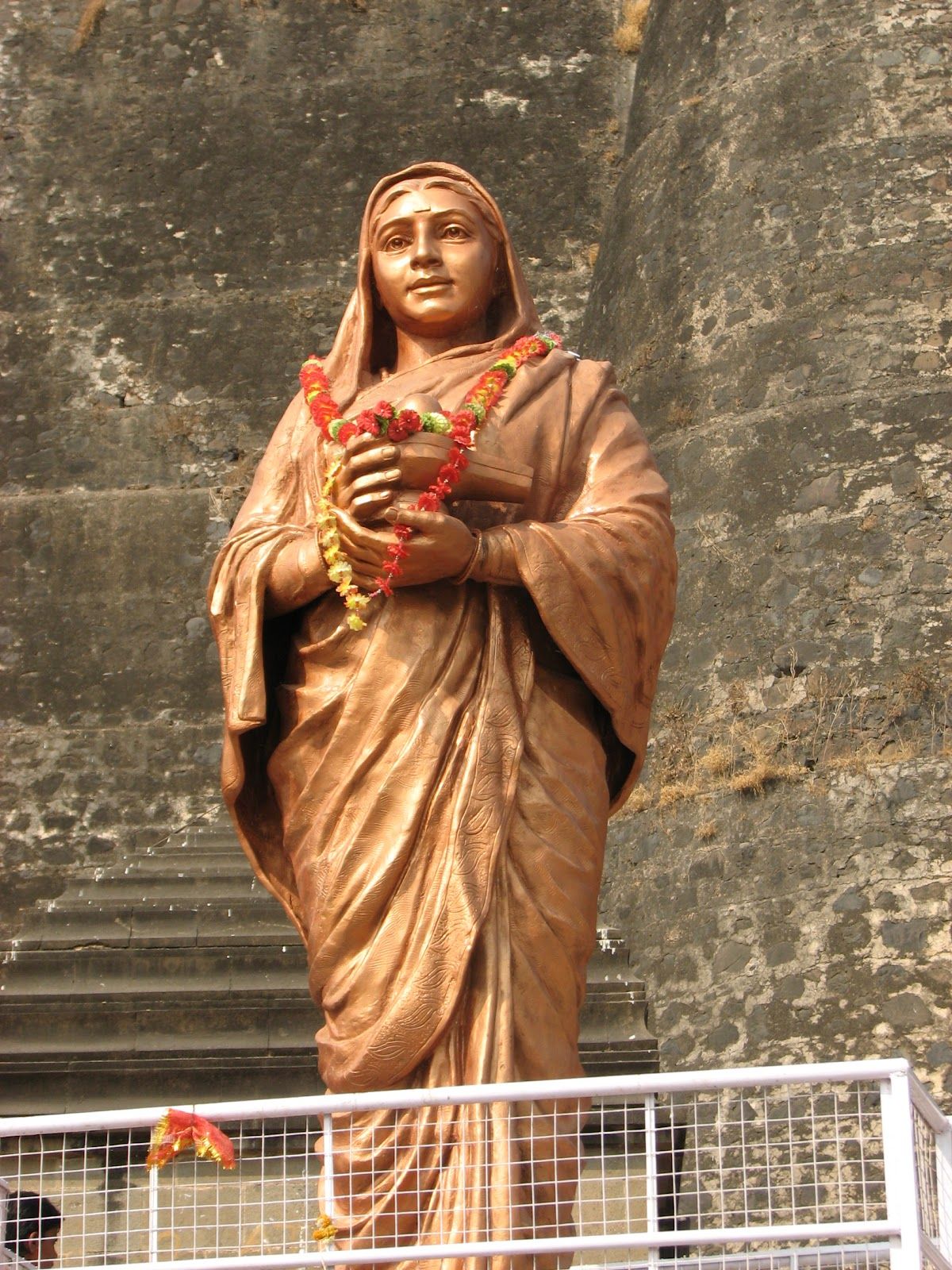 Bronze statue of Ahilyabai Holkar, garlanded, before a stone fort wall.